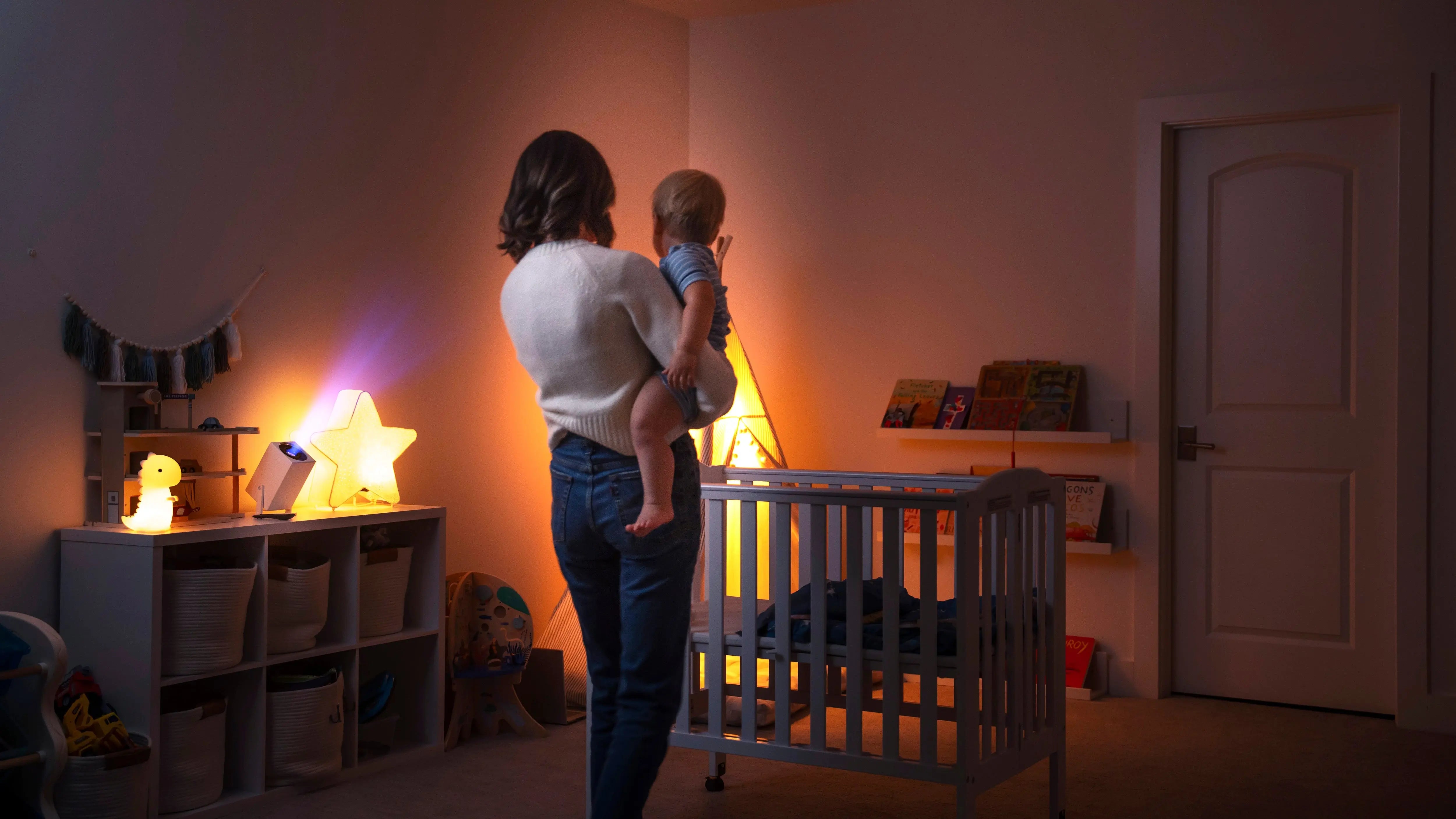 Mother holding a baby in a nursery, standing near a crib and a dresser with an Ozmotic Learning projector and star light.
