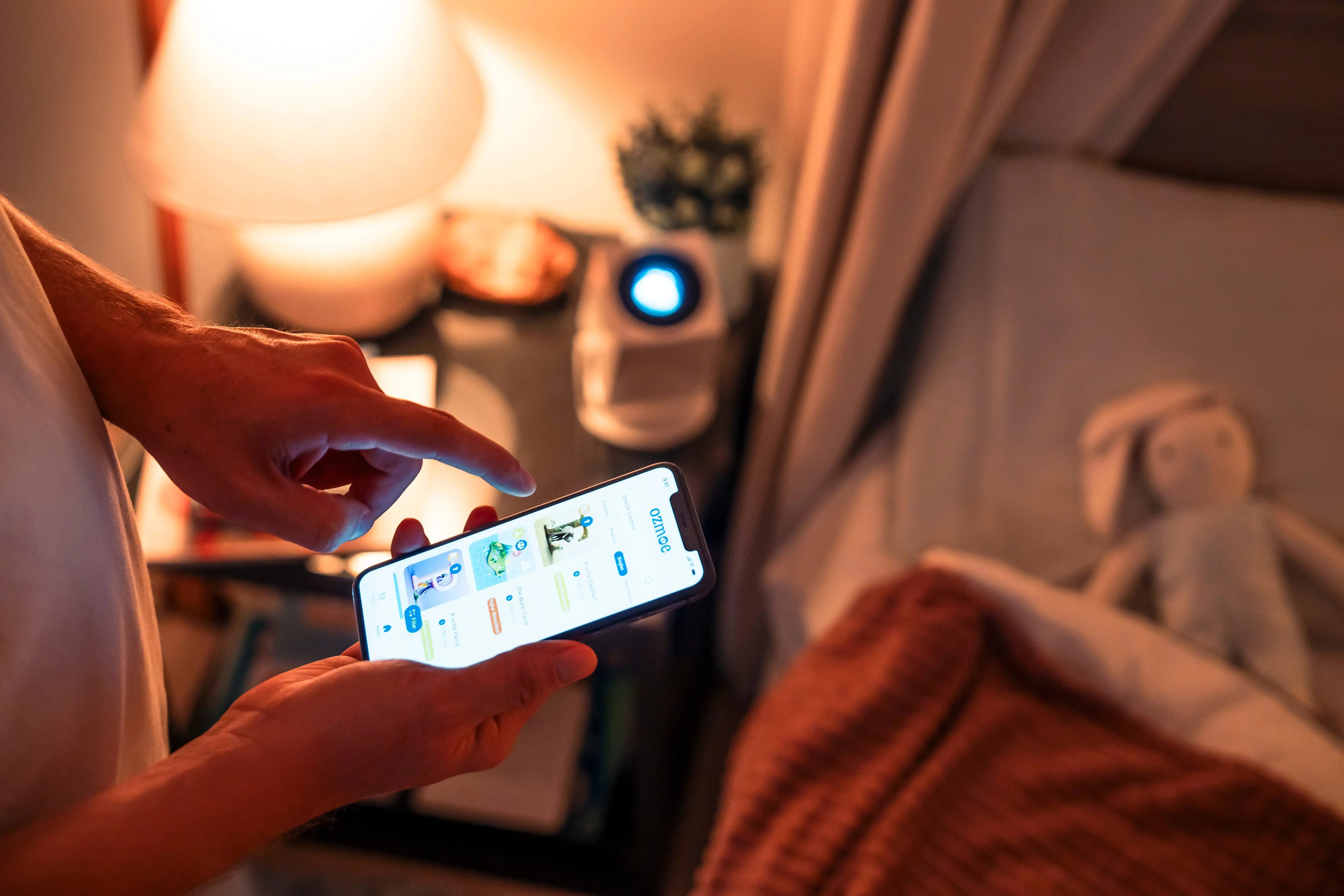 Parent using the Ozmotic Learning app on a smartphone to control the bedtime projector on a nightstand.