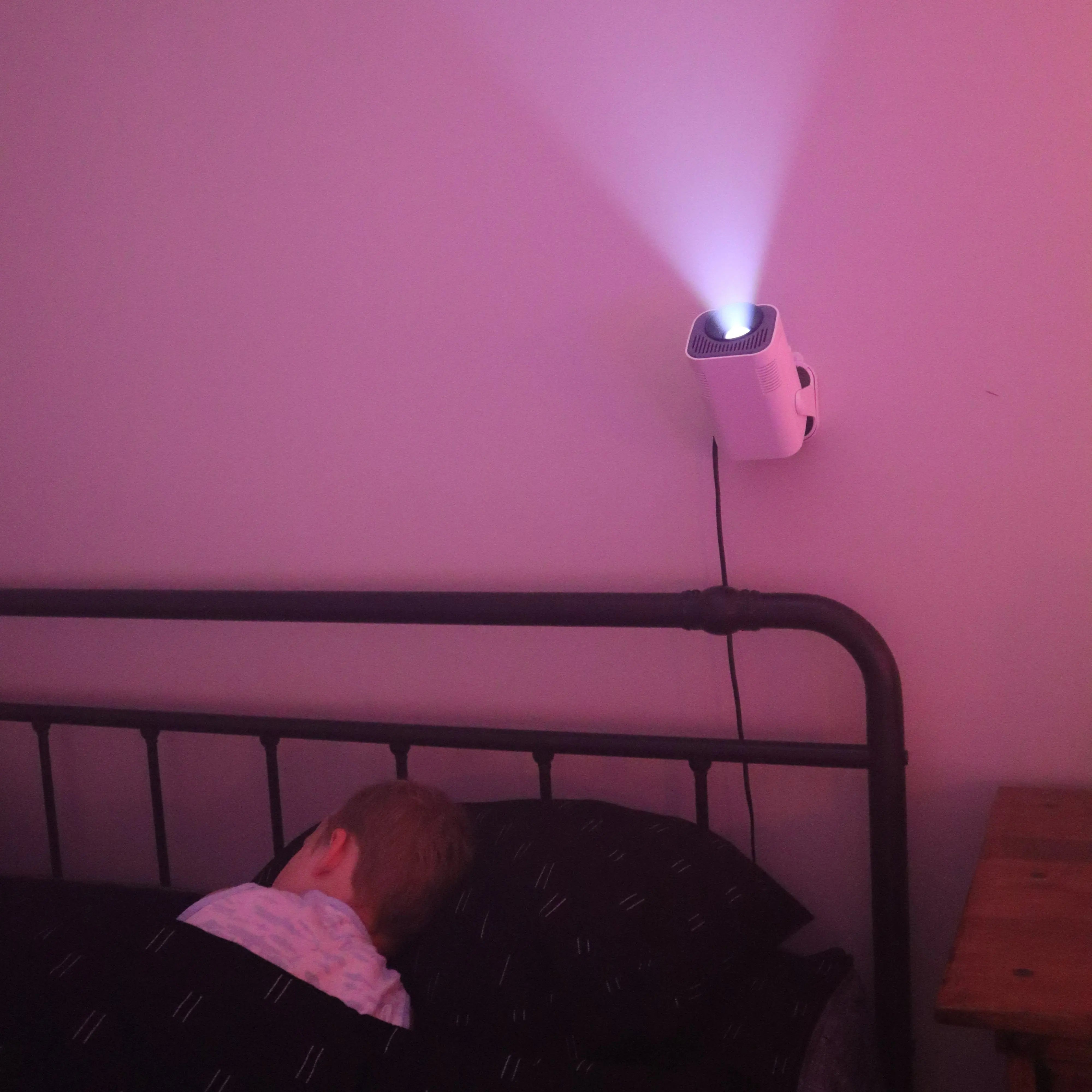 Child sleeping in a bed under an Ozmotic Learning projector mounted on the wall, casting a soft pink light.
