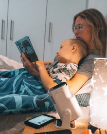 A heartwarming evening in bed: A mother, wearing glasses and with her blonde hair, is cuddled under a blue patterned blanket, holding a colorful open children's book. Her young child leans contentedly against her, looking up at the pages. On the adjacent table, a small white cylindrical Ozmotic Learning projector rests beside a smartphone and water bottle, casting a gentle light as they share a story.
